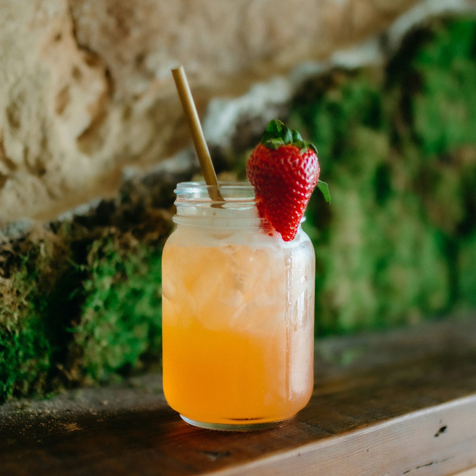 Das Bild zeigt einen Cocktail, der auf einem Holzbrett steht. Das Glas ist ein Marmeladenglas und der orange farbene Cockteil ist mit einer Erdbeere garniert. 